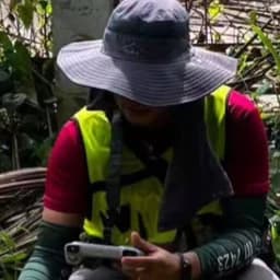 Jabez Joshua Flores agachado en un sitio de levantamiento topográfico, sosteniendo un controlador de dron