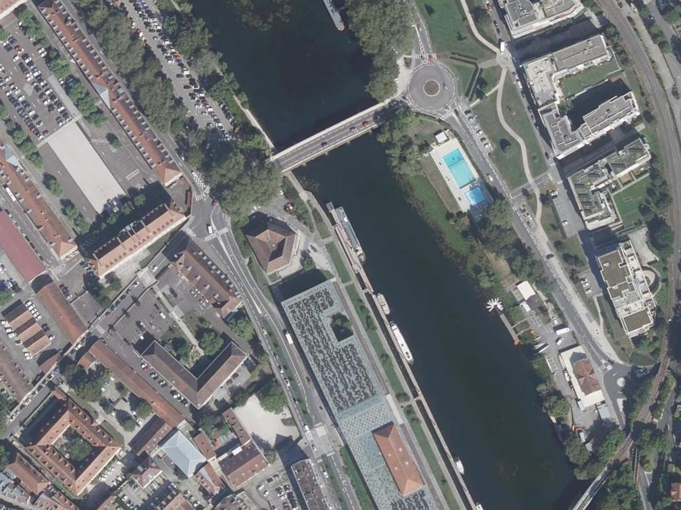 Aerial view of the Doubs through central Besançon, with dense old town on the left bank, quays, moored boats, a bridge and an open-air pool on the right bank.
