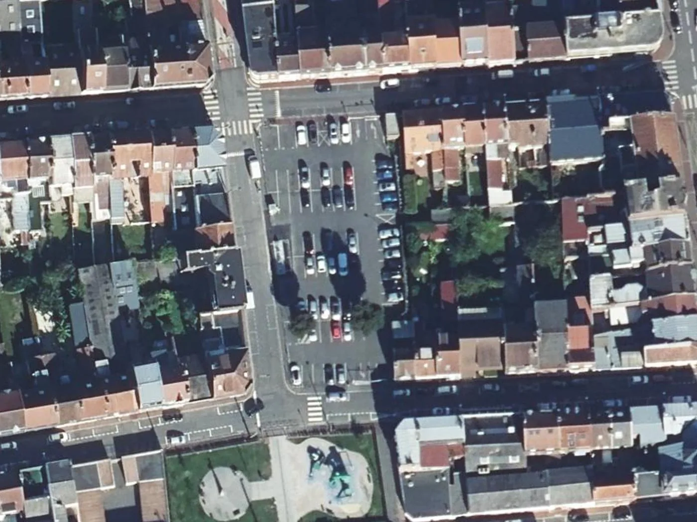 A surface car park in the middle of a block in the centre of Lens on the IGN orthophoto, about 160 m across, with terraced houses and their back gardens around it and a small square with a statue to the south.